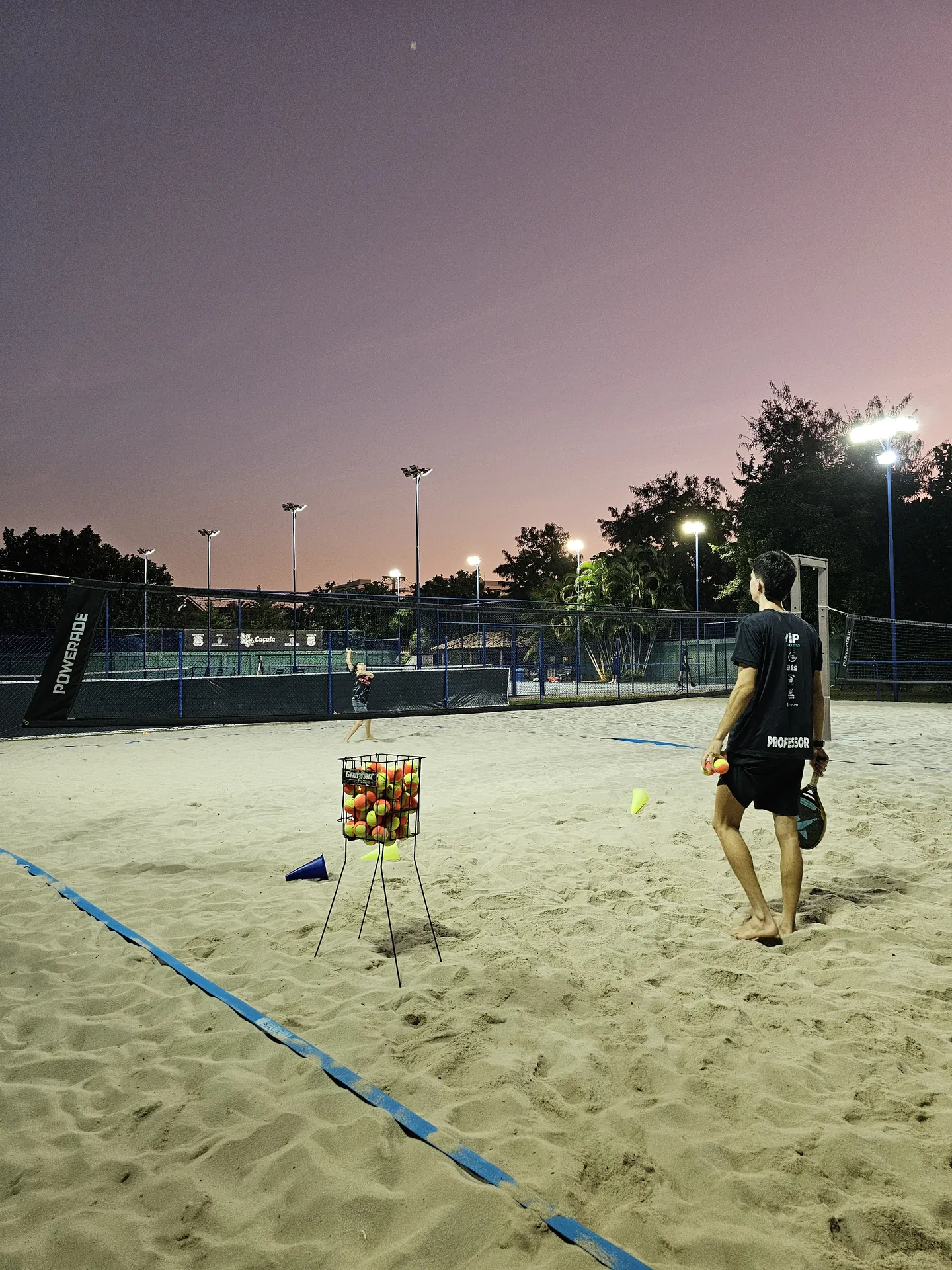 Treino de beach tennis no Instituto Tennis Route ao entardecer, com professor e cesto de bolas na quadra de areia